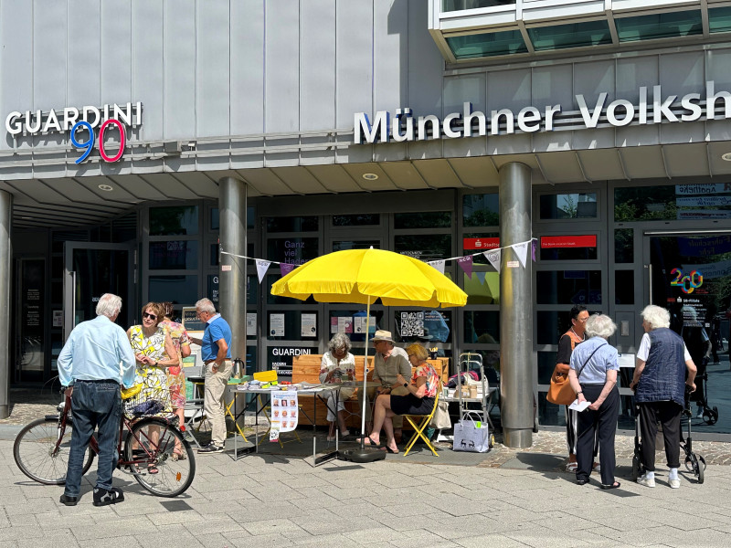unter einem großen gelben Sonnenschirm steht eine Holzbank, die Haderner MITMACH-Bank genannt wird
